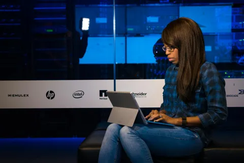 Young woman wearing glasses working on a laptop in a server room with technology company logos on the glass panel.