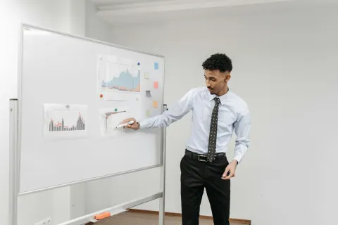 Man in formal attire presenting graphs and charts on a whiteboard during a business meeting.