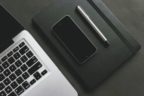 Laptop keyboard, smartphone on closed black notebook, and silver pen arranged on dark surface