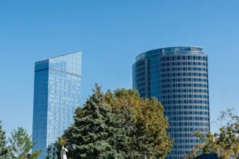 Modern glass skyscrapers behind lush green trees under a clear blue sky in an urban park setting