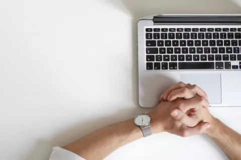 Person with folded hands wearing a wristwatch next to a laptop and a green plant on a white desk.