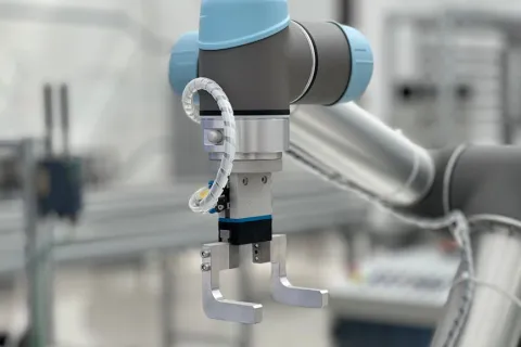 Close-up of a robotic arm with a metal gripper and blue protective covers in a manufacturing setting.