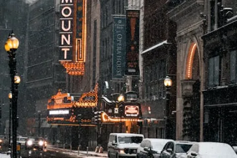 Snowfall on a city street with lit Paramount theater sign and parked cars covered in snow during evening.