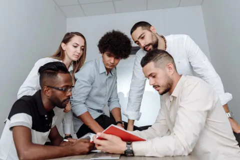 Diverse team of five young professionals gathered around a red notebook collaborating in a modern office.