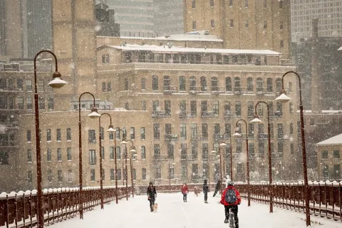 Snow falls on a city bridge with pedestrians and a cyclist in winter, surrounded by urban buildings.