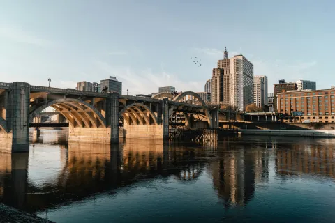 Sunlit city skyline and arched stone bridge reflecting over calm river water under a clear sky.