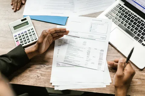 Person reviewing financial documents with calculator and laptop on wooden desk workspace.