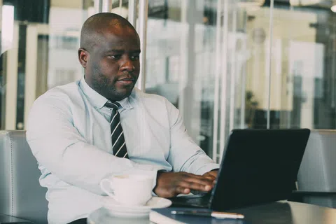 Professional man in shirt and tie working on laptop at cafe table with coffee cup and pen nearby