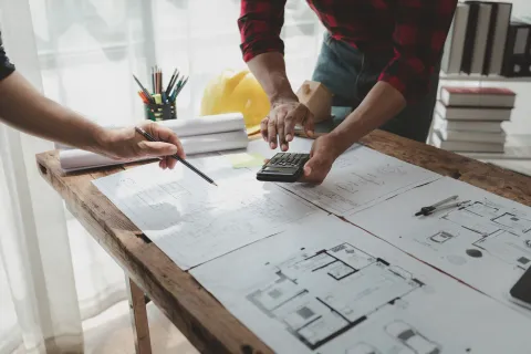 Two architects discussing building plans with a calculator, blueprints, and design tools on a wooden table.