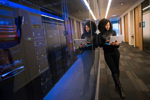 Woman in black outfit using a laptop in a server room with reflective glass and blue lights