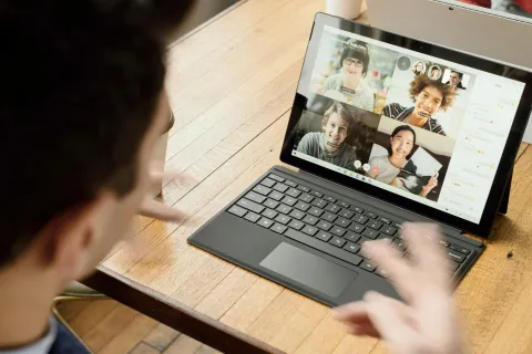 Person using laptop for video conference call with four people visible on screen, sitting at wooden table.