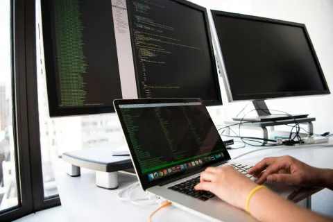 Person coding on a laptop with software code on dual desktop monitors in a bright office workspace.