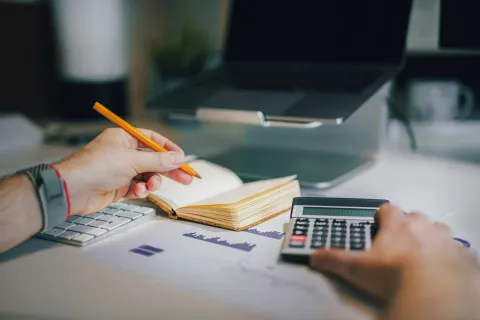 Person using calculator and writing notes on a notebook with a laptop and financial charts on desk.