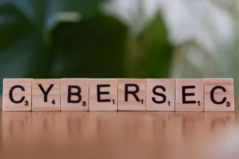Wooden Scrabble tiles spell out the word CYBERSEC on a wooden surface with a blurred green background