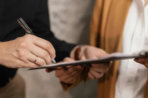 Two professionals in business attire reviewing and signing documents on a clipboard indoors.