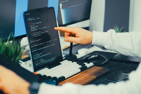Developer pointing at code on a tablet screen at a modern workspace with keyboard and monitors.