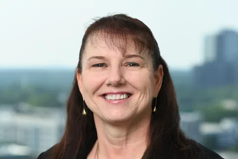 Smiling woman with dark hair wearing black blazer and green top with gold necklace and earrings, cityscape background.