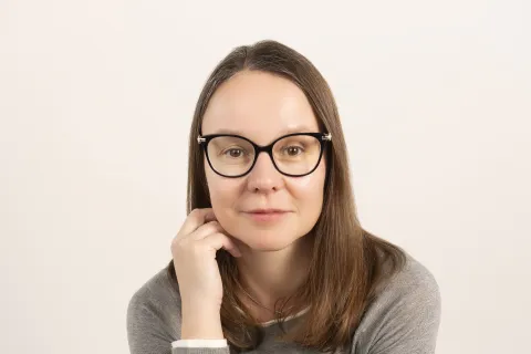 Woman with glasses wearing gray sweater and black pants sitting on a stool against a plain background