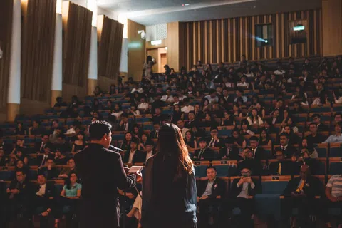 Speaker and woman addressing a large audience in a formal auditorium setting with attentive crowd listening.