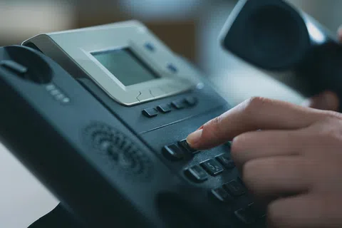 Close-up of a hand dialing buttons on a black office desk phone with a digital display.