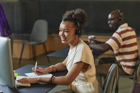 Smiling customer service representative with headset working at computer and taking notes in office