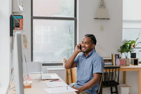 Man in blue shirt sitting at desk smiling while talking on phone in bright home office with laptop and books
