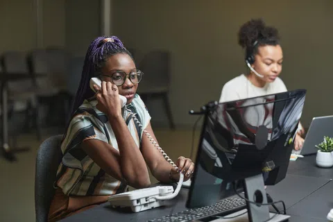 Two women working at office desks, one talking on a landline phone and the other using a headset and laptop.
