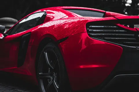 Close-up of a shiny red sports car rear with rain droplets and sleek tail light design under natural lighting.