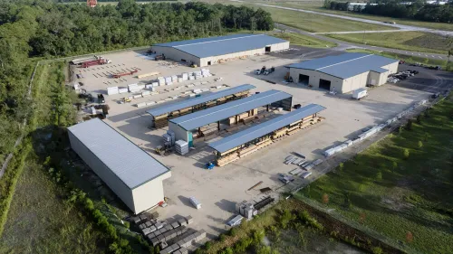 Aerial view of a lumber yard with multiple storage buildings and covered wood storage racks surrounded by greenery.