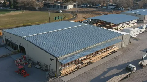 Aerial view of a large industrial warehouse with metal roofing and outdoor lumber storage under a covered area.