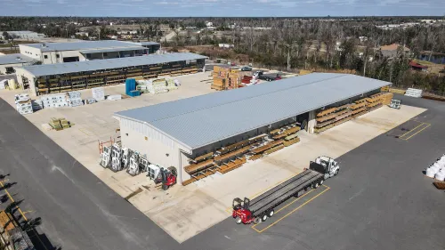 Aerial view of a large building materials warehouse with stacks of lumber and trucks in a spacious yard under a clear sky.
