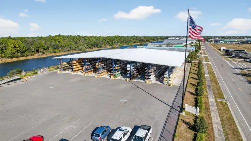 Lumber yard with stacked wooden planks and forklift under a covered storage area on a clear day