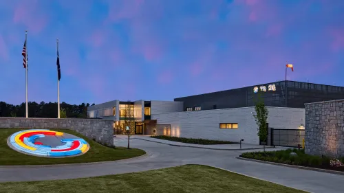 Modern two-story medical building with ambulance, cars, greenery, and partly cloudy sky overhead.