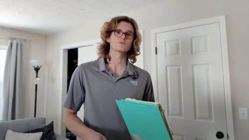 Young man with glasses holding teal clipboard standing in a bright living room with gray sofa and closed doors.
