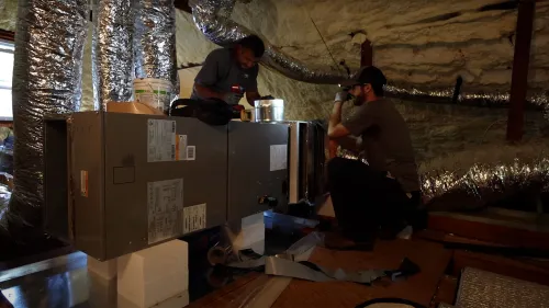 Two technicians install and inspect HVAC equipment in an insulated attic space with ductwork.