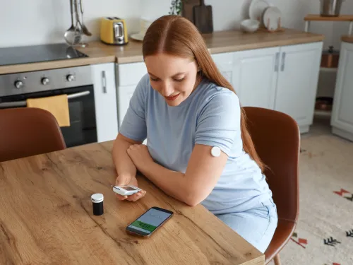 Person with glucose monitor sensor on arm checking data on smartphone screen indoors