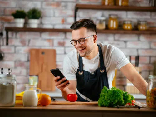 Man in apron smiling while looking at phone in kitchen surrounded by fresh vegetables and ingredients