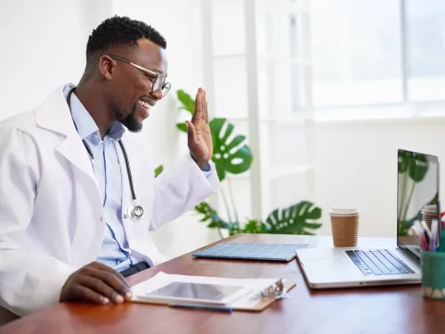 Smiling doctor in white coat waving during a video call on laptop in a bright office setting.