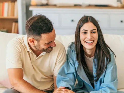 Smiling couple sitting on couch holding hands and enjoying a happy moment together indoors.