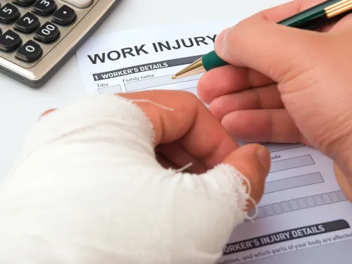 Person with a bandaged hand filling out a work injury report form with a pen near a calculator.