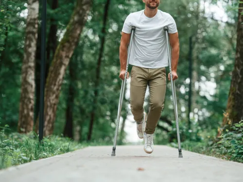 Man with crutches walking on a forest path surrounded by trees and greenery on a sunny day.