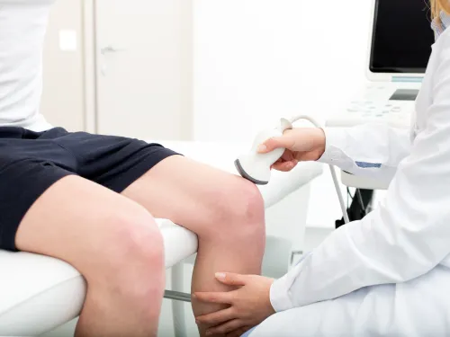 Doctor performing ultrasound scan on patient's knee using medical imaging device in clinic.