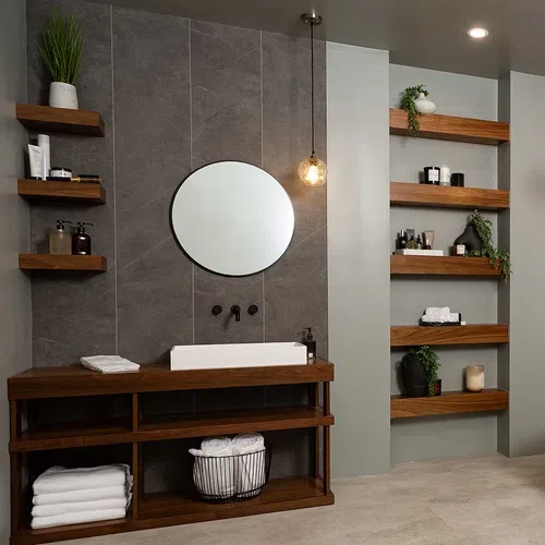 Modern bathroom with wood shelves, round mirror, white sink, gray tiles, and decorative plants and towels.