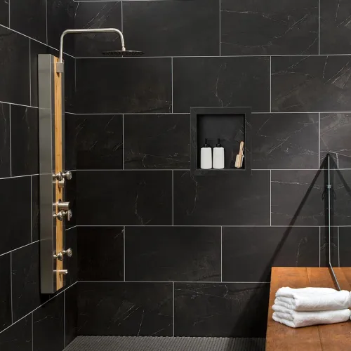 Modern black tiled shower with stainless steel shower panel, built-in shelf with bottles, and wooden bench with towels