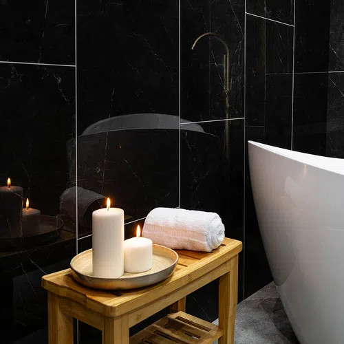 Wooden bench with lit candles and a rolled towel next to a white bathtub in a black marble bathroom.