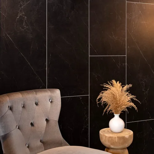 Beige tufted chair beside a wooden stool with a white vase and dried pampas grass against black marble wall.