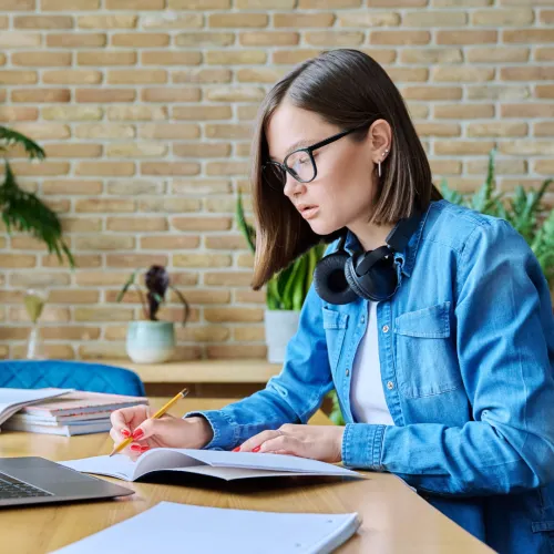 Young woman in glasses and denim jacket writing in notebook with laptop and headphones on wooden desk