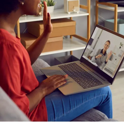 Woman in red shirt video chatting on laptop, waving to a person on the screen in a home setting.
