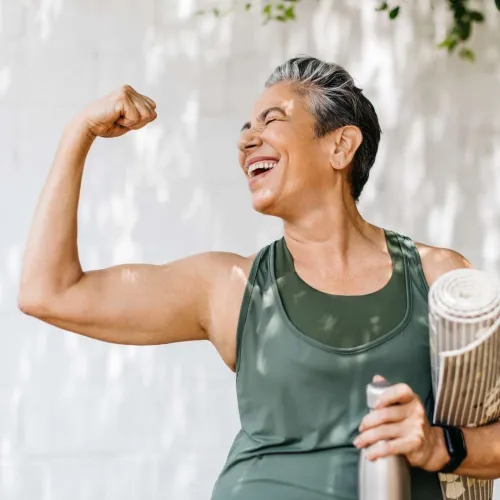 Mature woman in green athletic wear flexes arm and smiles while holding a yoga mat and water bottle.