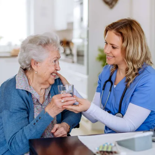Nurse in blue scrubs helping elderly woman hold a glass of water, both smiling in a home setting.